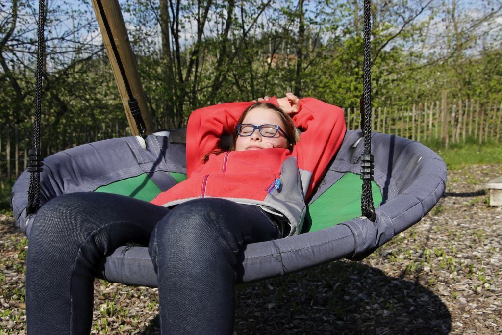 Kind liegt entspannt mit geschlossenen Augen auf der Nestschaukel in der Sonne auf dem Spielplatz des Ferienhofs